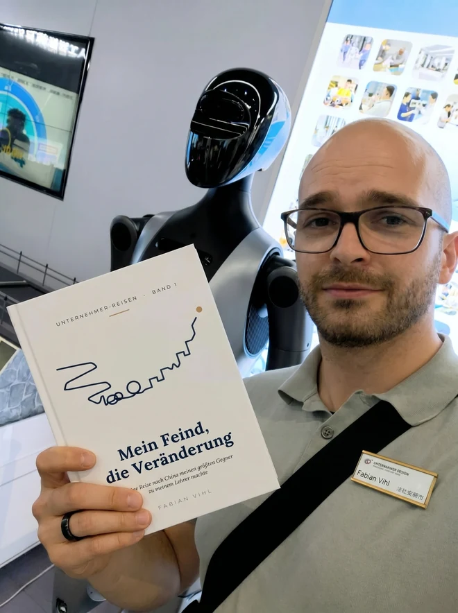 Fabian Vihl hält das gedruckte Buch in die Kamera, hinter ihm ein humanoider Roboter auf einem Messestand